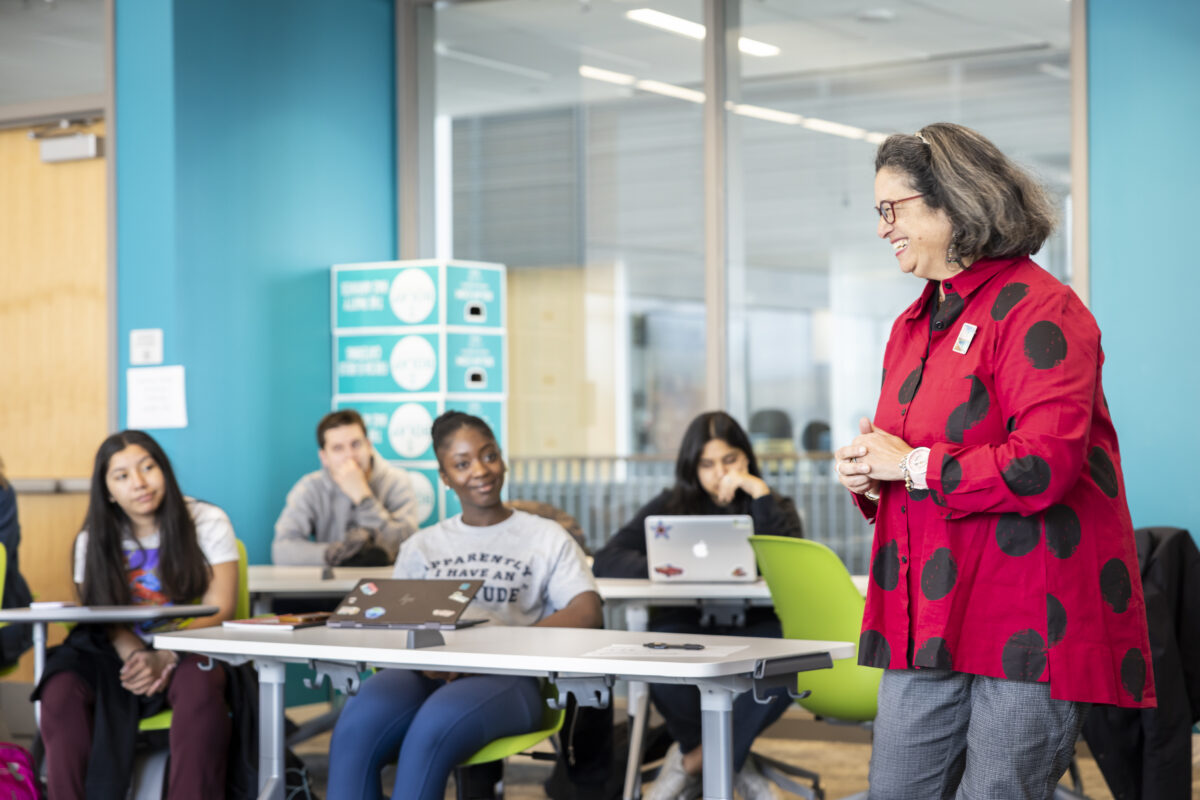 A professor from Schar School stands in the middle of a classroom teaching while four students are sitting at a table looking at the professor.
